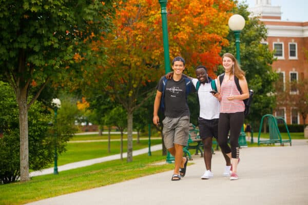 Two male students and one female student walk together while smiling on the Castleton campus during peak foliage