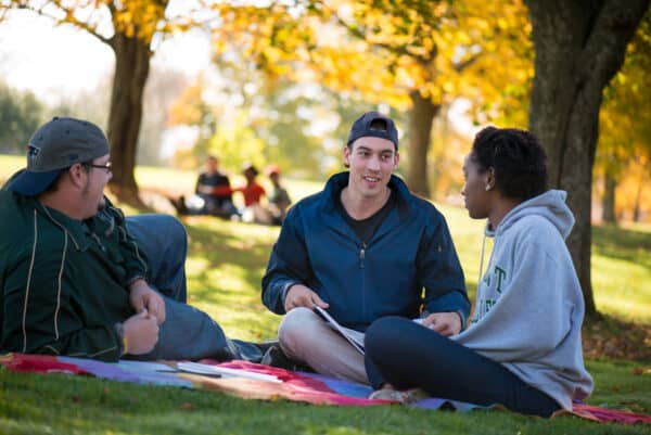 Two male students and one female student sit outside and chat under the trees
