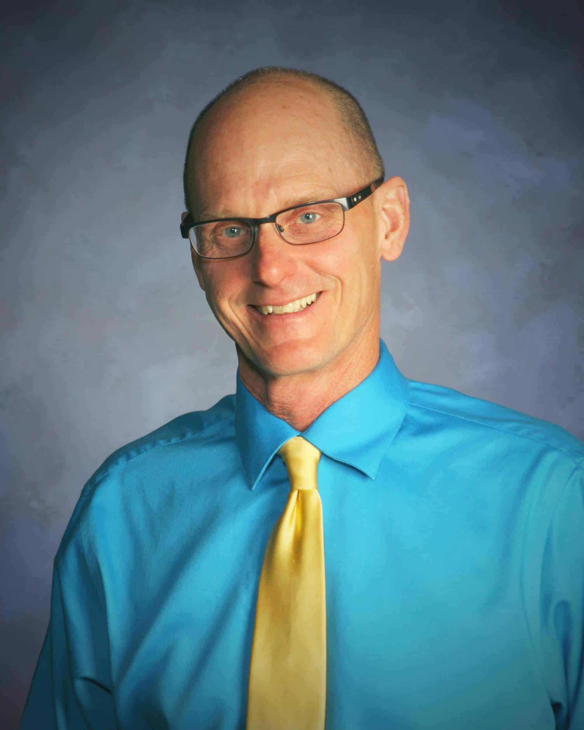 A bald man in glasses with a blue shirt and yellow tie smiles at the camera.