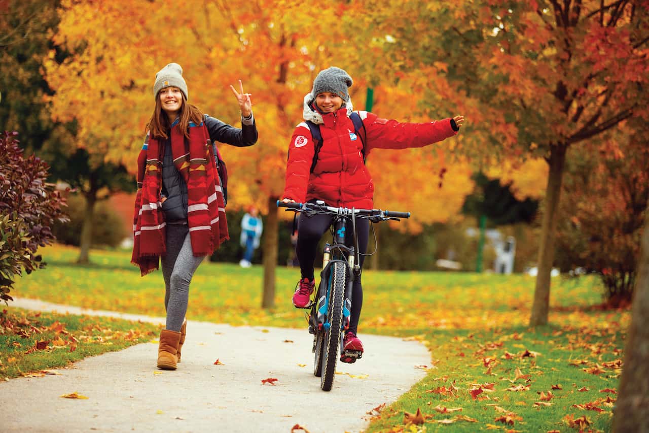 Two female students wave to the camera while walking and biking around the Castleton campus