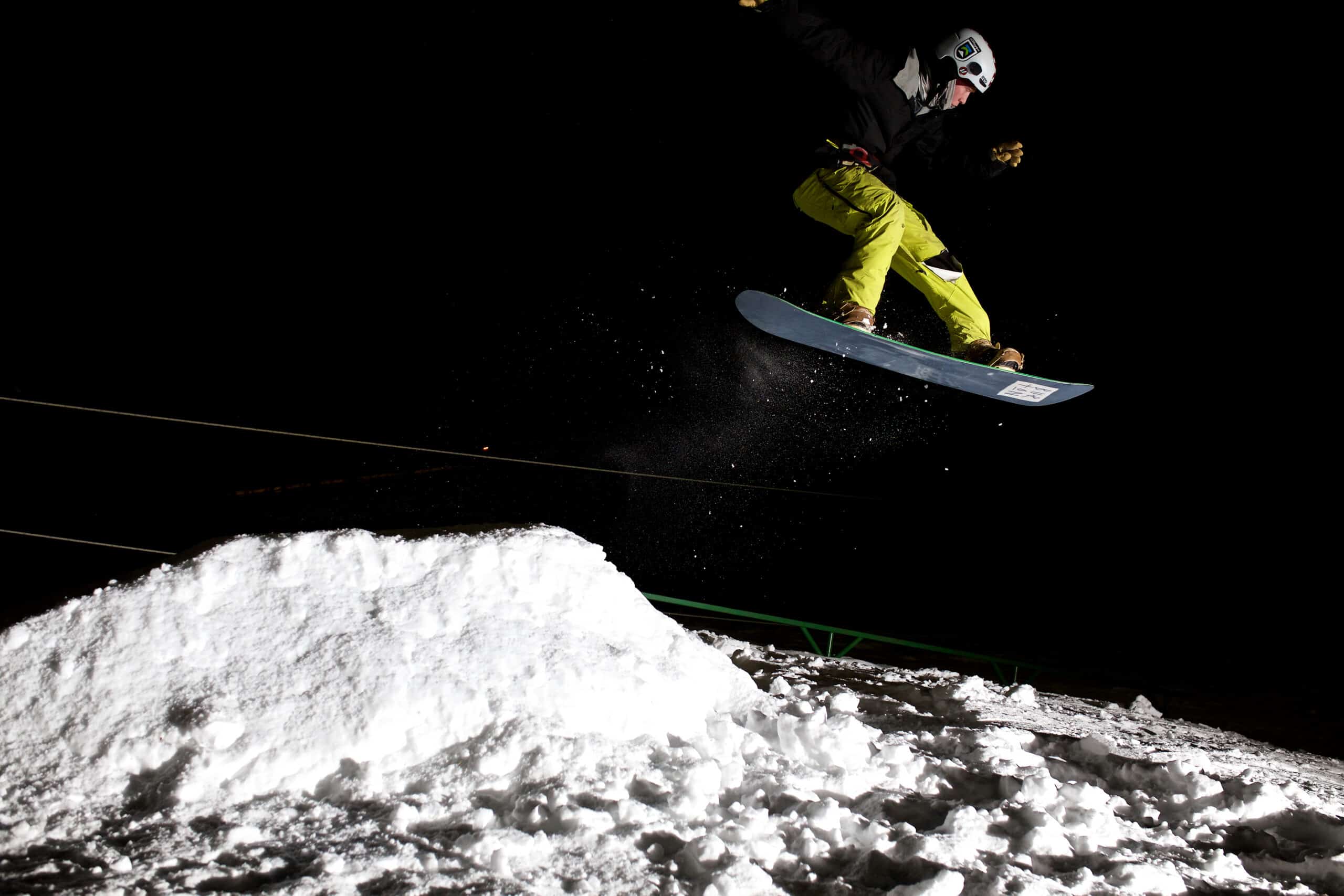 A snowboarder with yellow pants jumping high in the air after hitting a snow ramp.