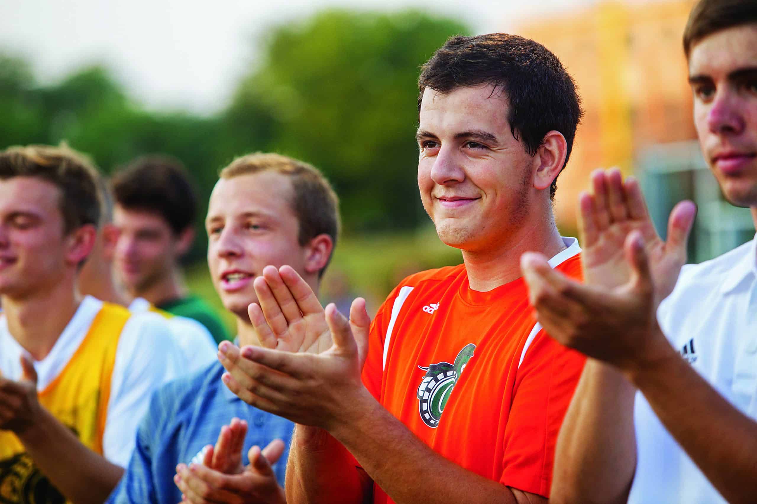 A brown haired young man in an orange shirt claps his hands in a crowd of other young men.