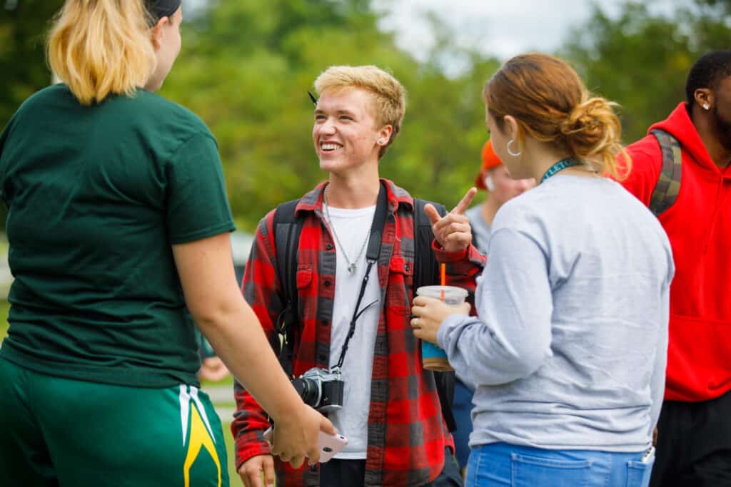 A young blond haired male student is smiling a laughing with two girls with their backs turned. He is wearing a red flannel shirt and a camera around his neck.