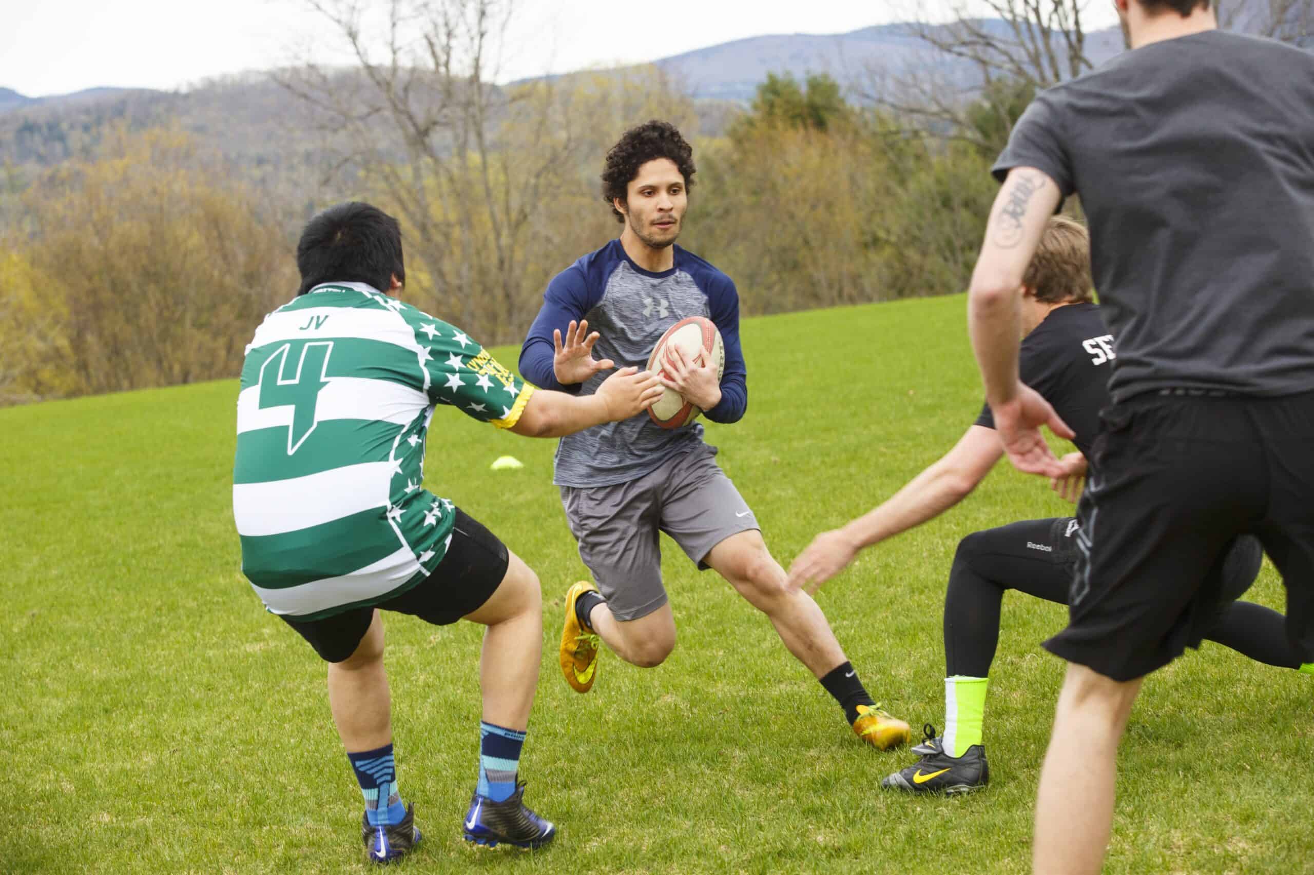 A young dark haired man holding a rugby ball runs towards a group of other men trying to stop him.