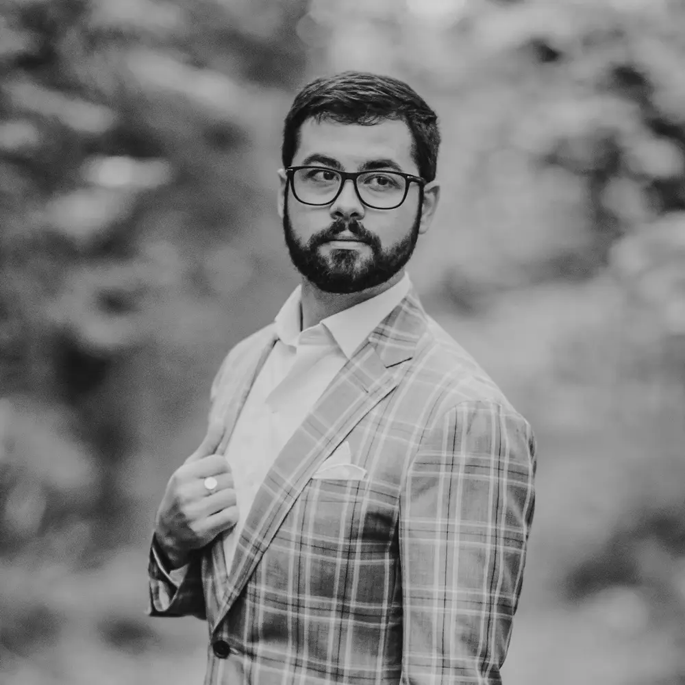A black and white photo of a young man with a beard and glasses wearing a plaid suit.