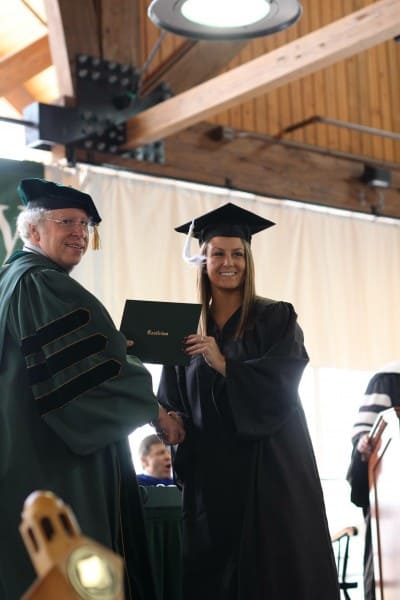 A photo of Ali Jones, a young woman with long blonde hair smiling in a black graduation robe. She is reaching with her left hand to receive her diploma