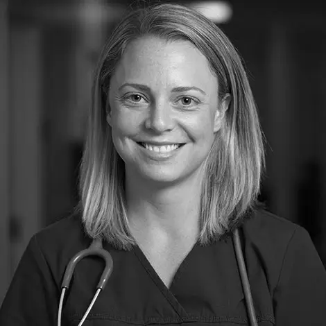 A black and white photo of a blond woman wearing scrubs and a stethoscope smiling at the camera. 