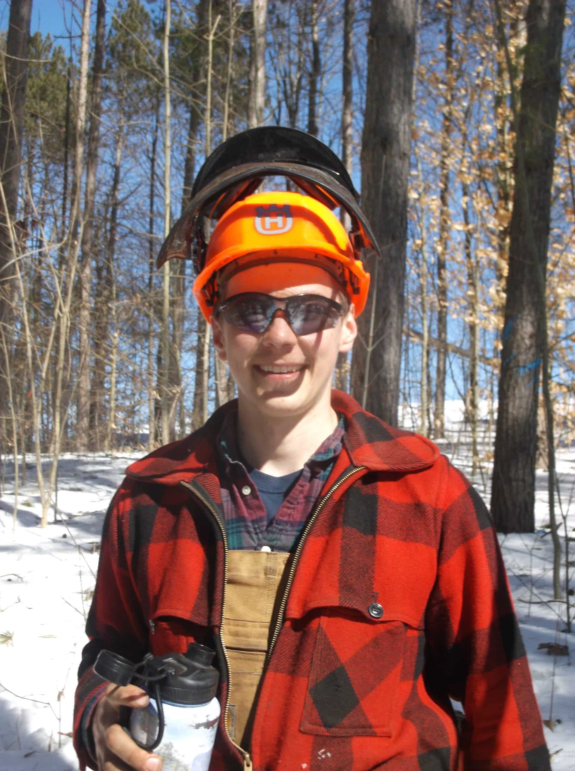 A young man standing in the snow with a coffee smiles at the camera while wearing an orange helmet and red plaid jacket.