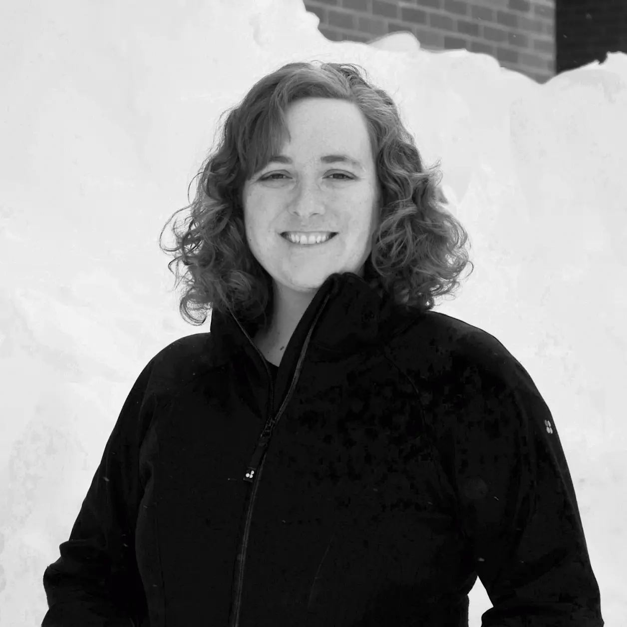 A black and white photo of a woman standing in the snow in a black jacket smiling at the camera.