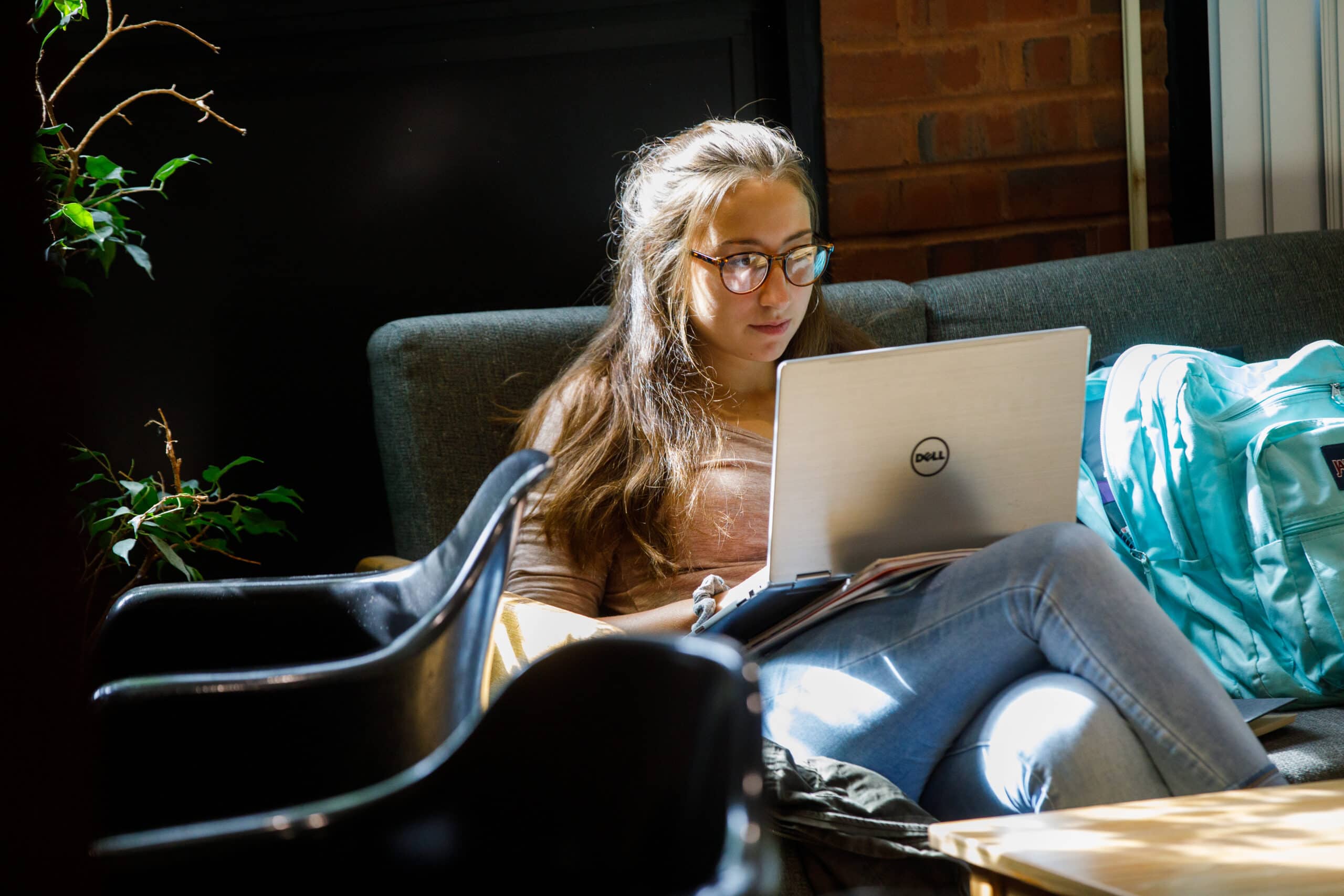 A girl with glasses sits on a couch with her laptop on her lap.