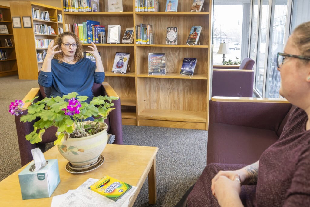 Two women sitting across from each other at a small table in a library. One is wearing a blue shirt and gesturing with her hands while the other leans in and listens.