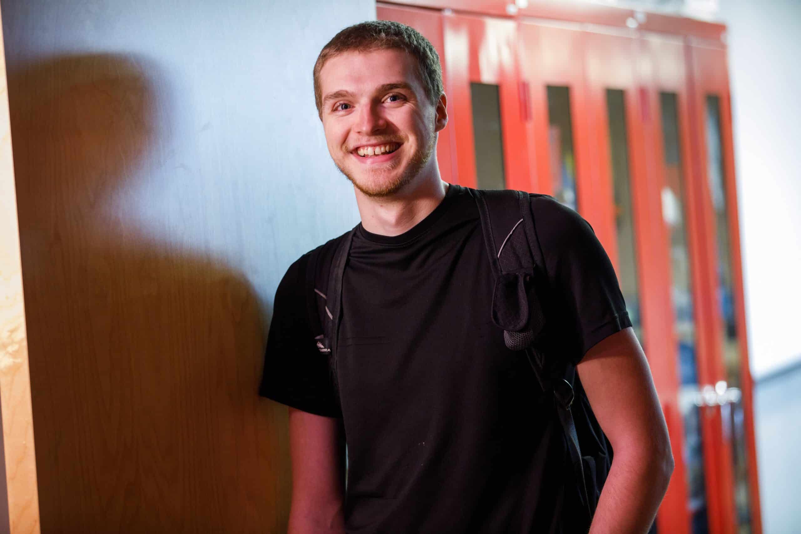 A young man with short hair in a black t-shirt smiles at the camera.