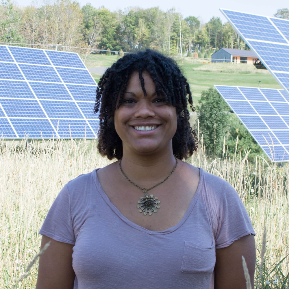 A woman with short curly hair smiles at the camera with solar panels in the background.
