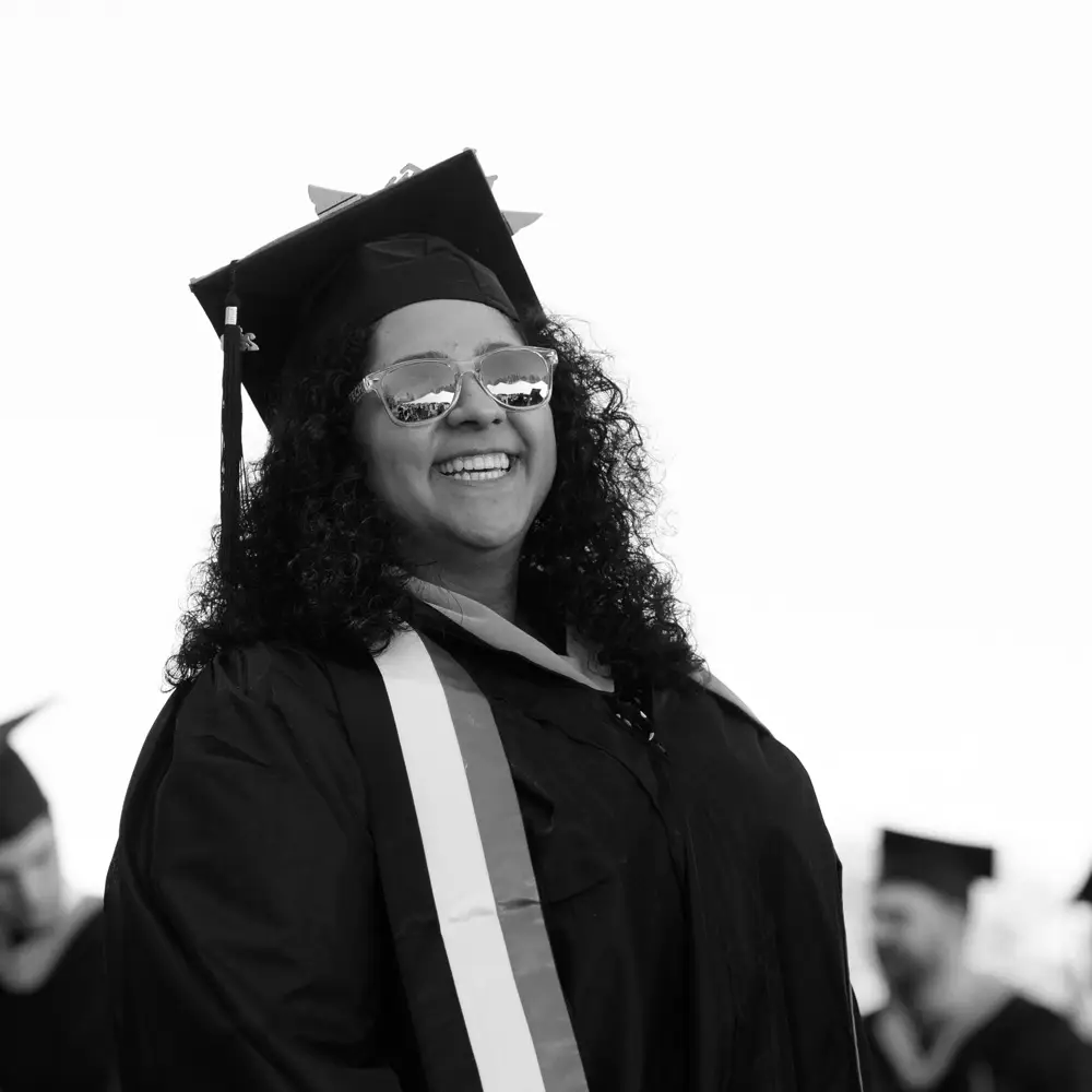 A black and white photo of a woman grinning in graduation regalia and sunglasses.