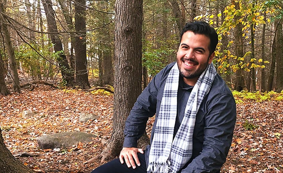 A dark haired man with a beard smiles at the camera while sitting in the woods wearing a scarf.