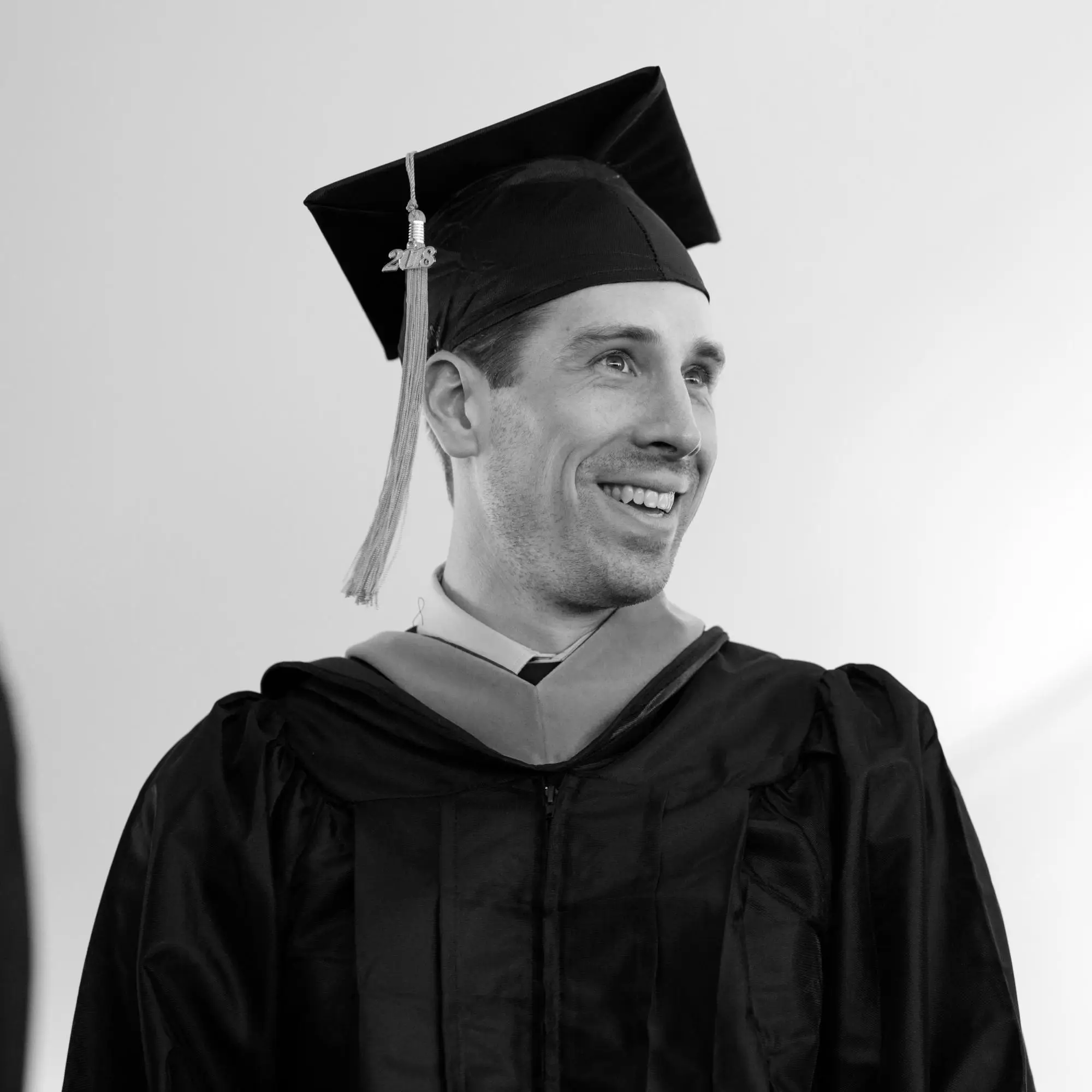 A black and white photo of a young man wearing graduation regalia smiling to the side.