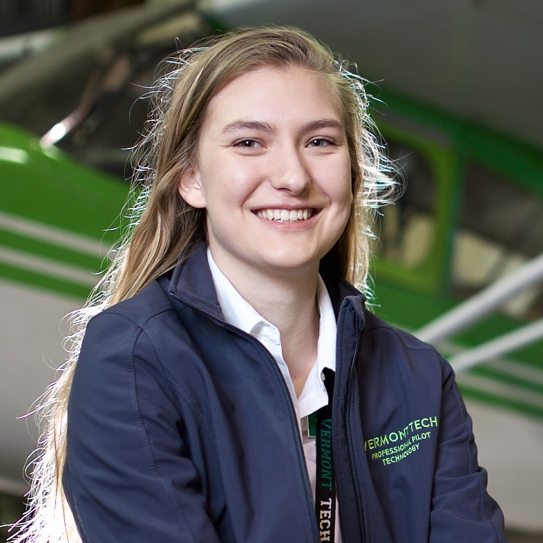 A photo of Elisabeth Hoehn, a young woman with long blonde hair smiling in front of a green and white airplane