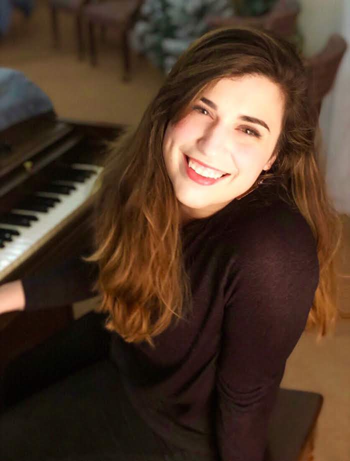 A photo of Gabby Fecher, a young woman with long brunette hair smiling sitting at a piano