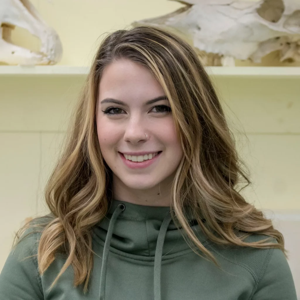 A photo of Great Khunly, a young woman with long brunette hair with blonde highlights wearing a green sweater, smiling and standing in front of a yellow wall with a shelf