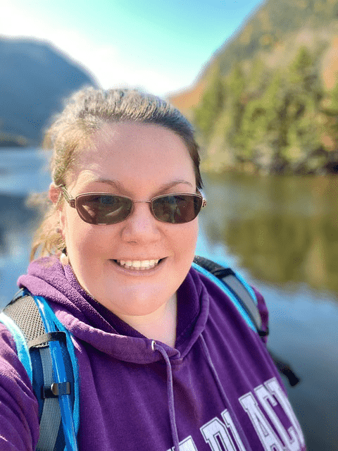 A woman in a purple sweatshirt and sunglasses takes a selfie with mountains and lake in the background.