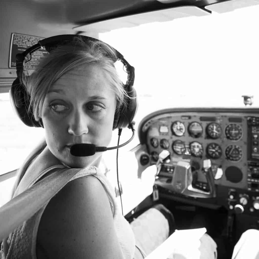 A black and white photo of Jamie Heiam, a young woman wearing a communication headset with her head turned over her right shoulder. She is seated in the pilots seat, steering an airplane