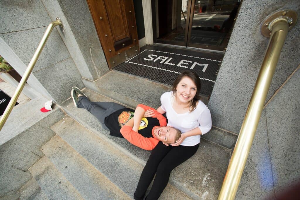 A photo of John and Olivia McDonough on concrete steps at the entrance of a building. Olivia is sitting in a white shirt and black pants with John laying in a orange shirt and denim pantson his back with his head in her lap
