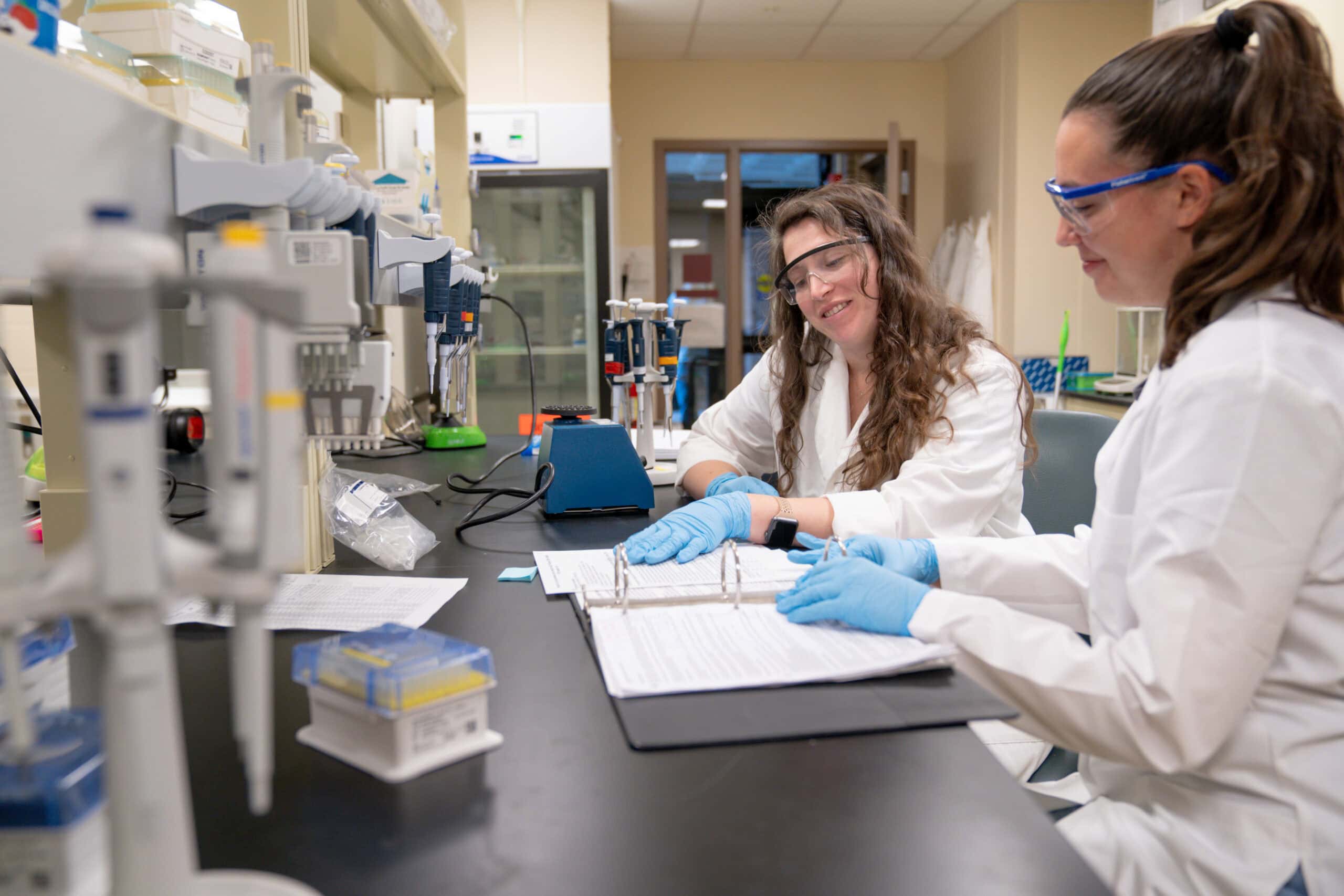 Two students wearing lab coats and eye protection working in a biology lab