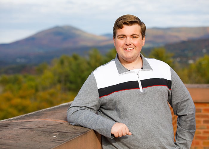 A young man in a striped polo leans against a brick balcony and smiles at the camera, mountains are in the background.