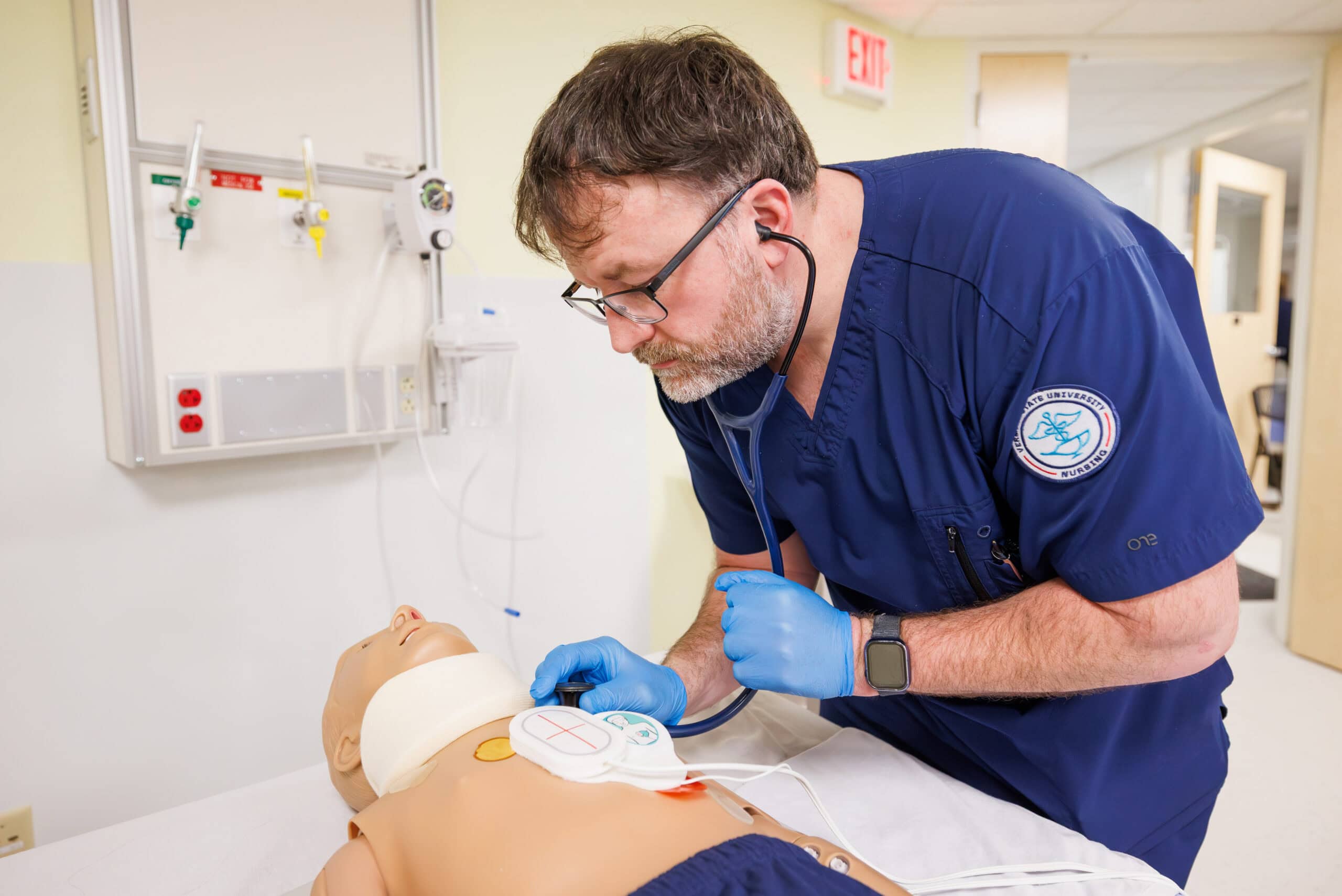 A male student wearing blue scrubs leaning over a training dummy in a nursing lab