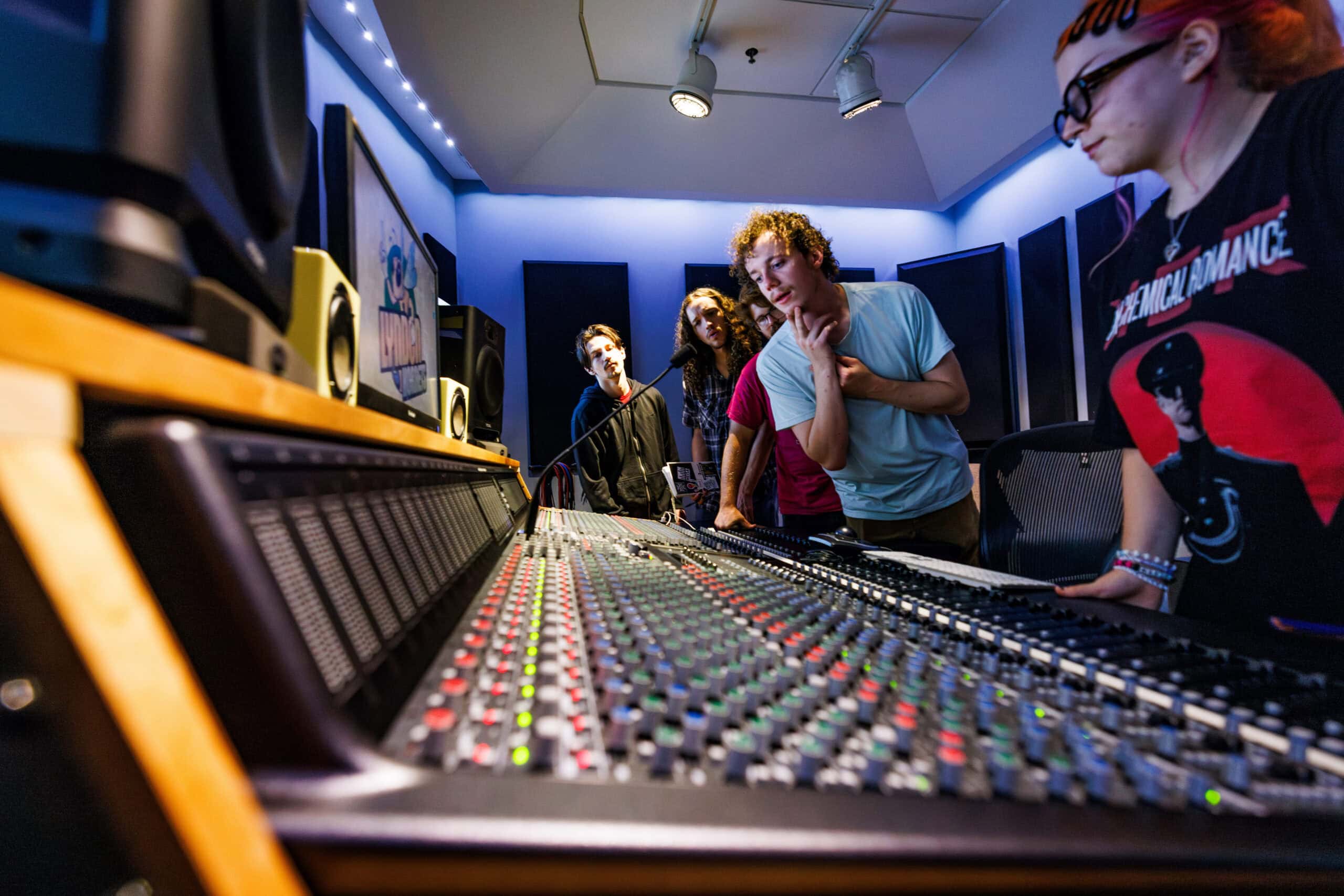 Students looking over an audio recording console while one of them speaks into a microphone