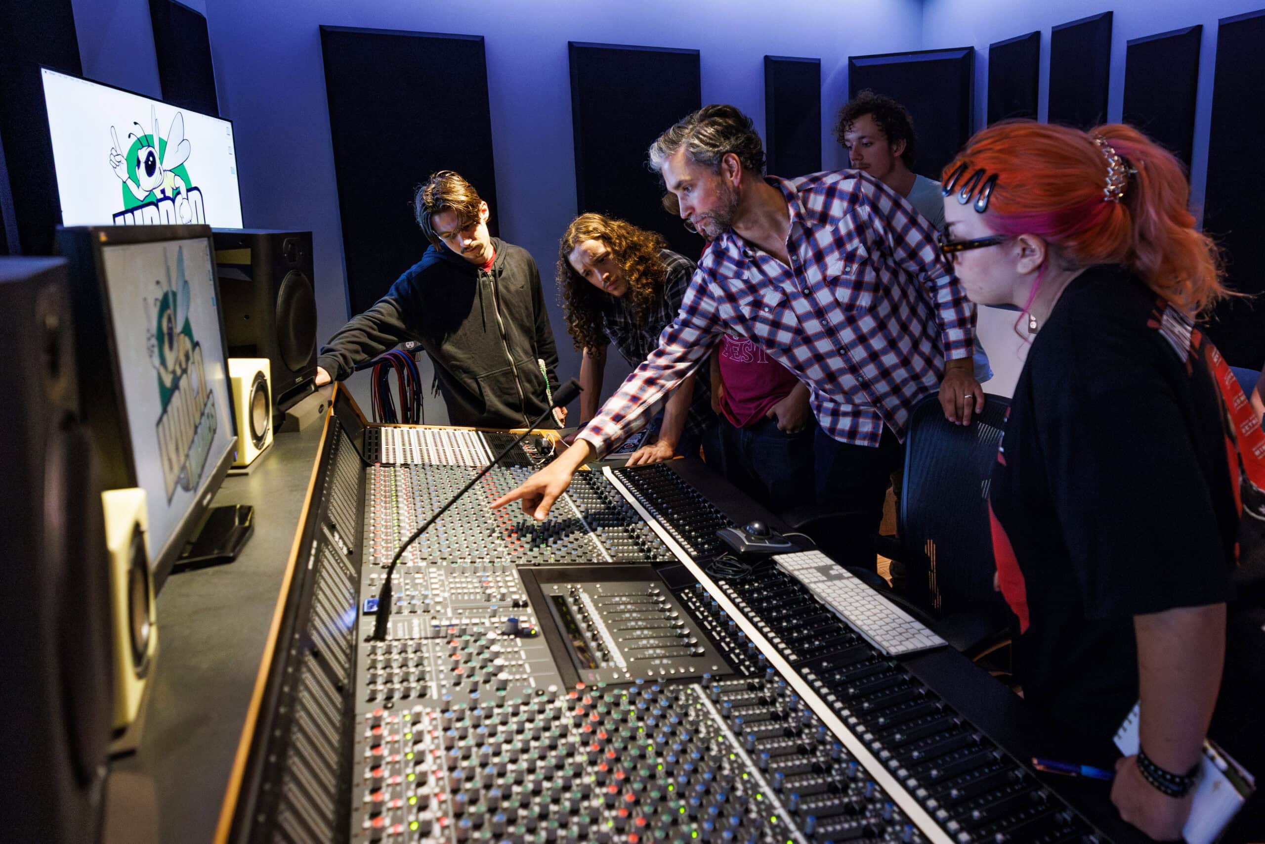A faculty member pointing at a a slider on an audio production console while students look on