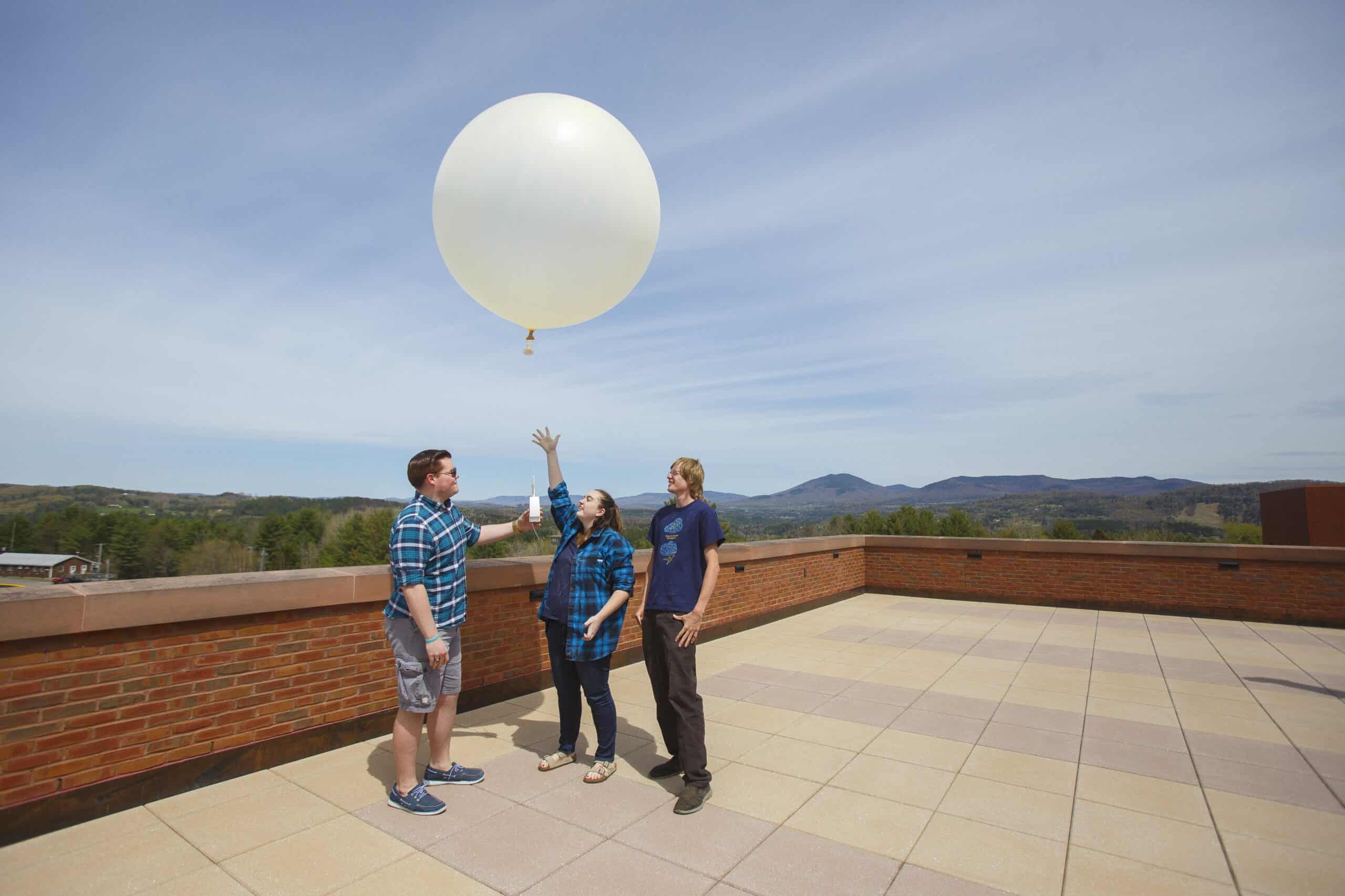 Three students stand in a circle on a rooftop releasing a giant white balloon.