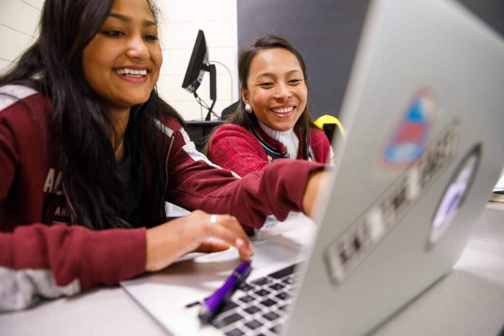 Two girls smiling and pointing at a laptop.