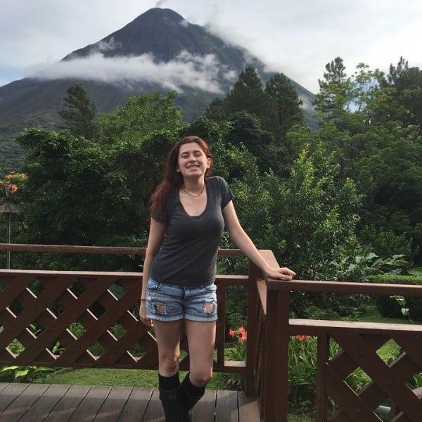 A dark haired girl leans against a railing with a large mountain in the background.