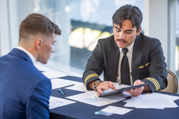 A man in a pilots uniform pointing at a tablet while speaking with a male student wearing a blue suit