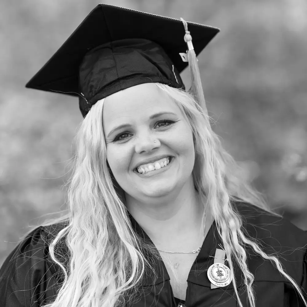 A black and white photo of a blond woman in a graduation cap and gown smiling at the camera.