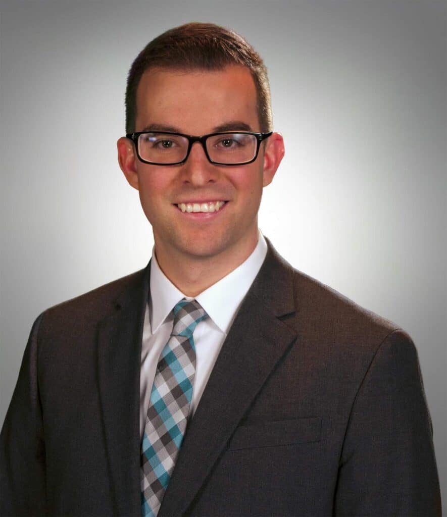 A photo of Tyler Cadorette, a young man with short brunette hair and rectangular glasses wearing a suit and smiling in front of a white background 