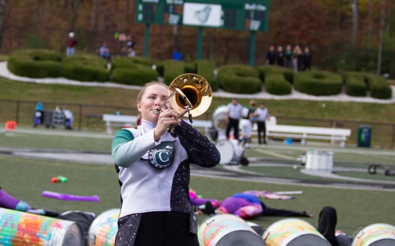 A woman in a cheerleading uniform leans back clutching a trumpet to her mouth in a stadium.