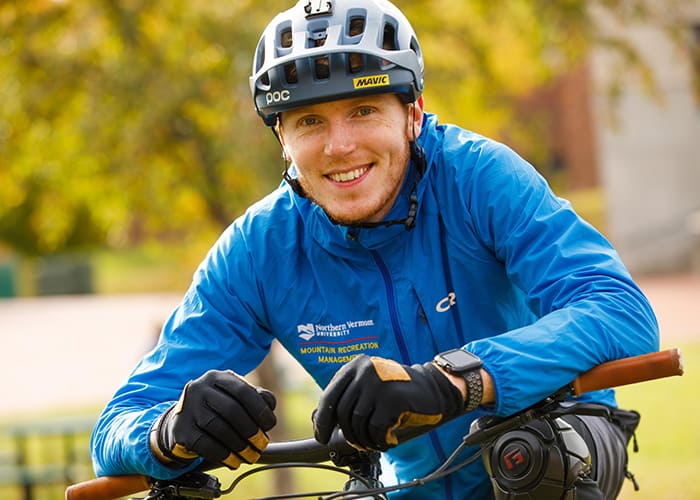 A young man leans over a bike smiling at the camera wearing a helmet and blue rain jacket.