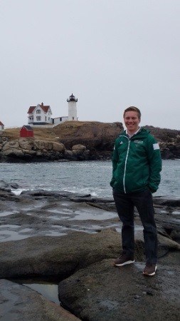 A photo of a young blond guy with short hair posing in front of a the coast with a lighthouse in the background.