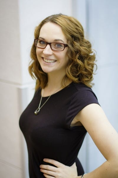 A young woman with long curly hair and glasses wears a black dress and smiles at the camera with her hands on her hips looking over her shoulder.