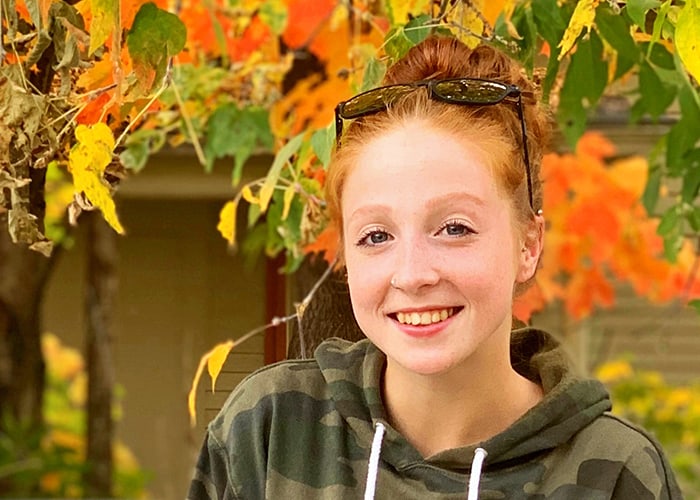 A red headed girl smiles for a portrait. She is wearing a camo hoodie and has sunglasses on her head.