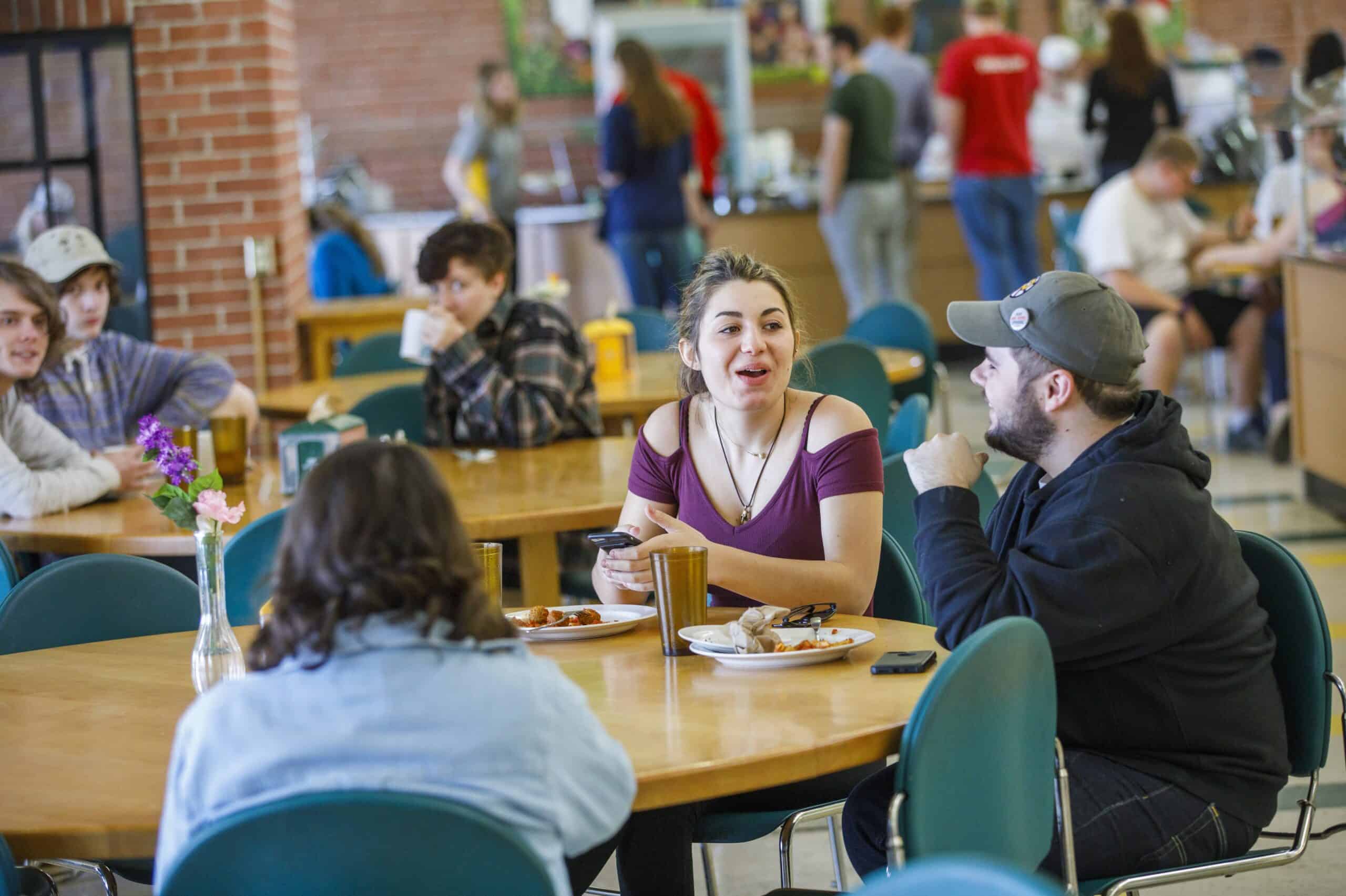 A girl is mid laugh as she's chatting with two friends at a table. They are in a cafeteria and there is a crowd of people in the background.