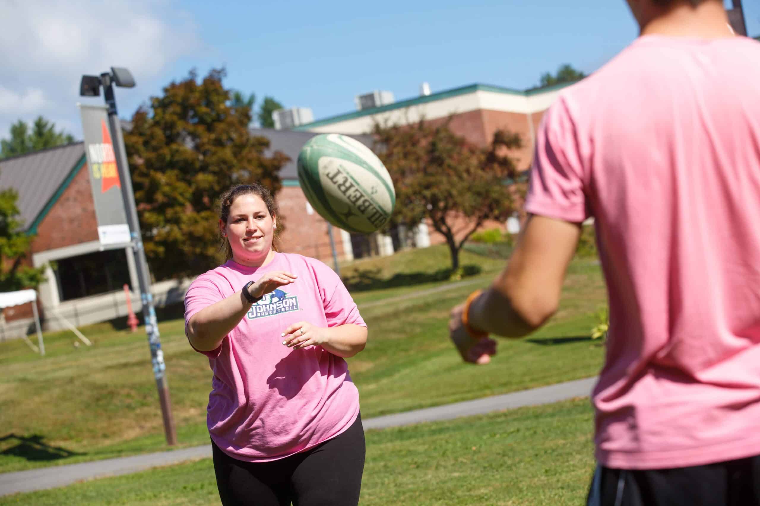 A long haired young woman in a pink shirt throws a rugby ball towards another person who's back is turned. They are outside and it's a sunny day.