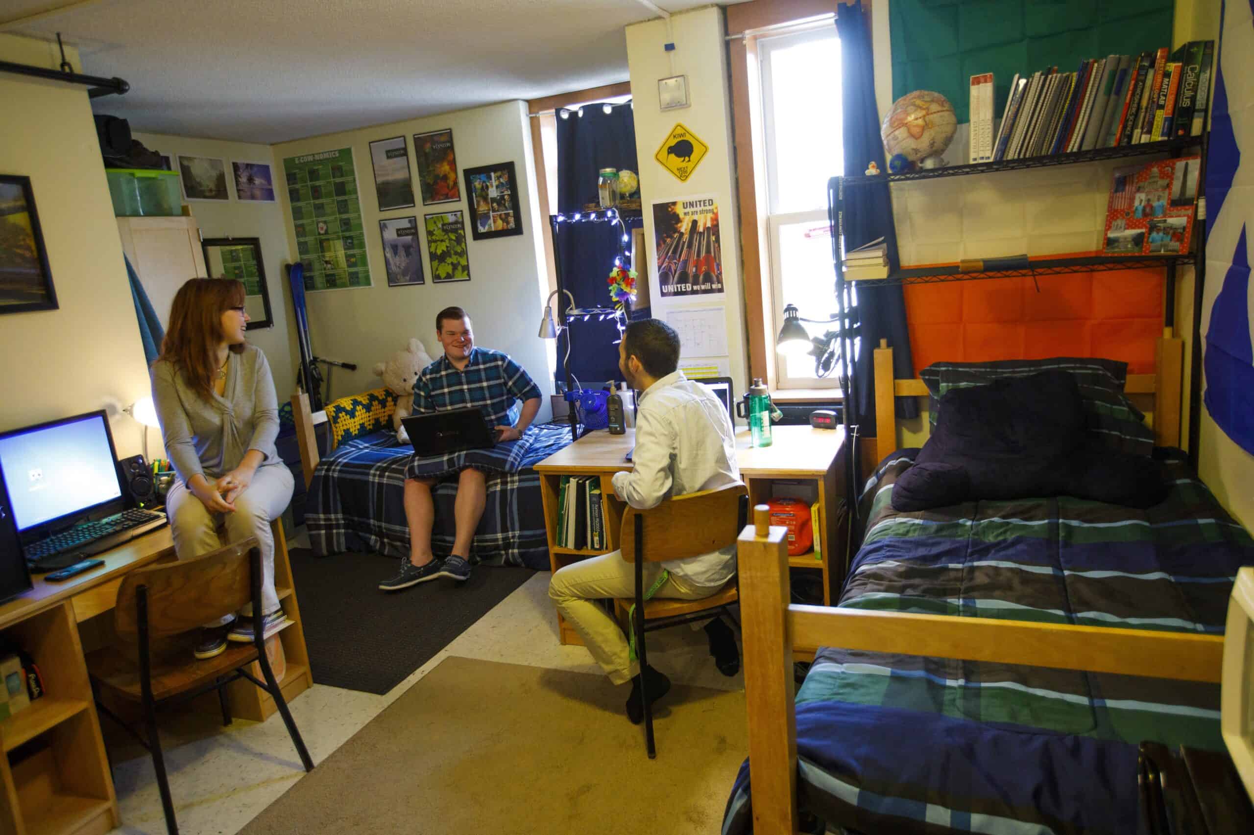 Two young guys and one girl are sitting in a dorm room having a conversation. The dorm room has two beds and two desks.