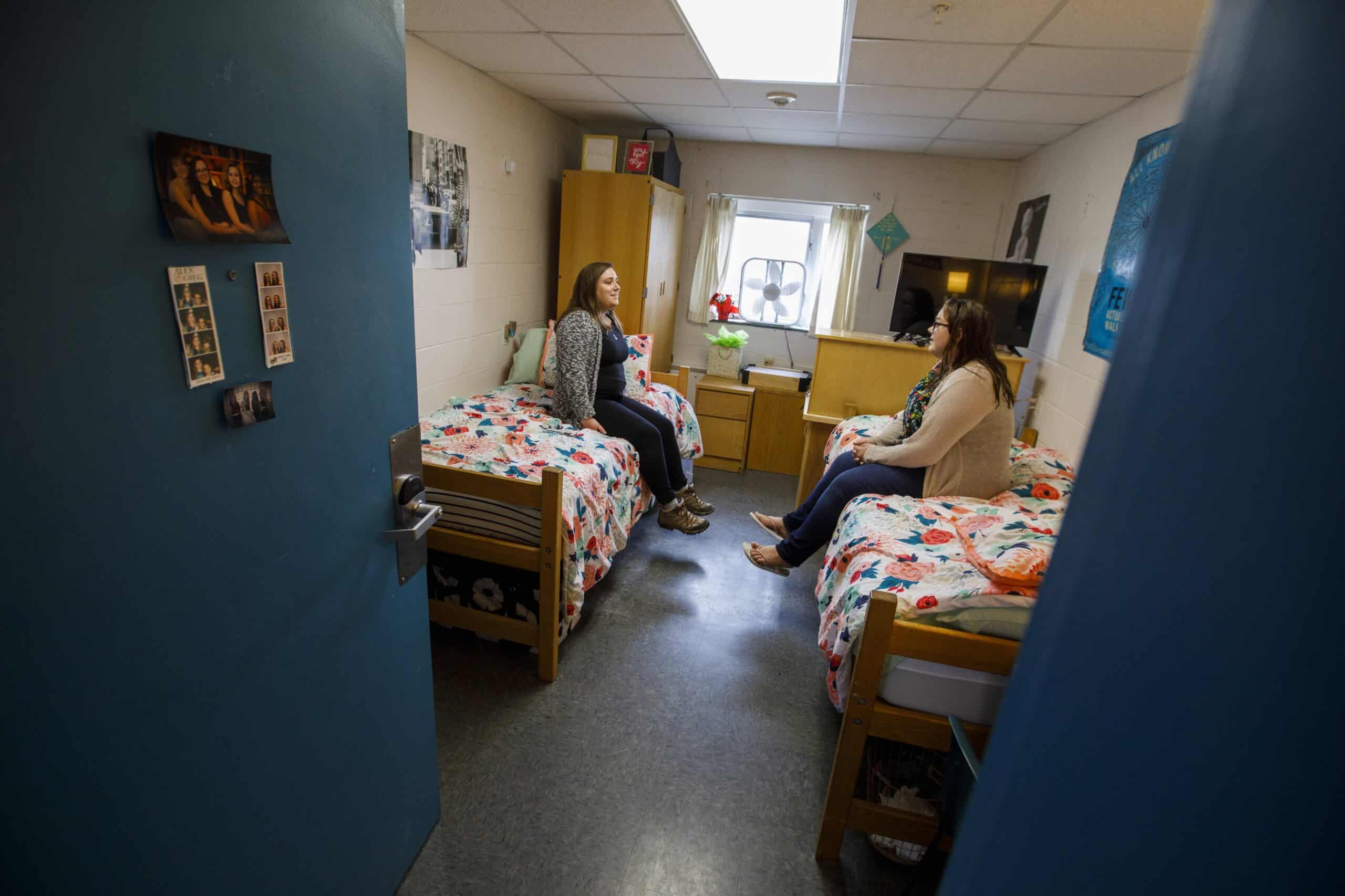 A dorm room with two beds on either side of the room. There is a girl on each bed facing each other and talking, their feet are dangling off the beds.