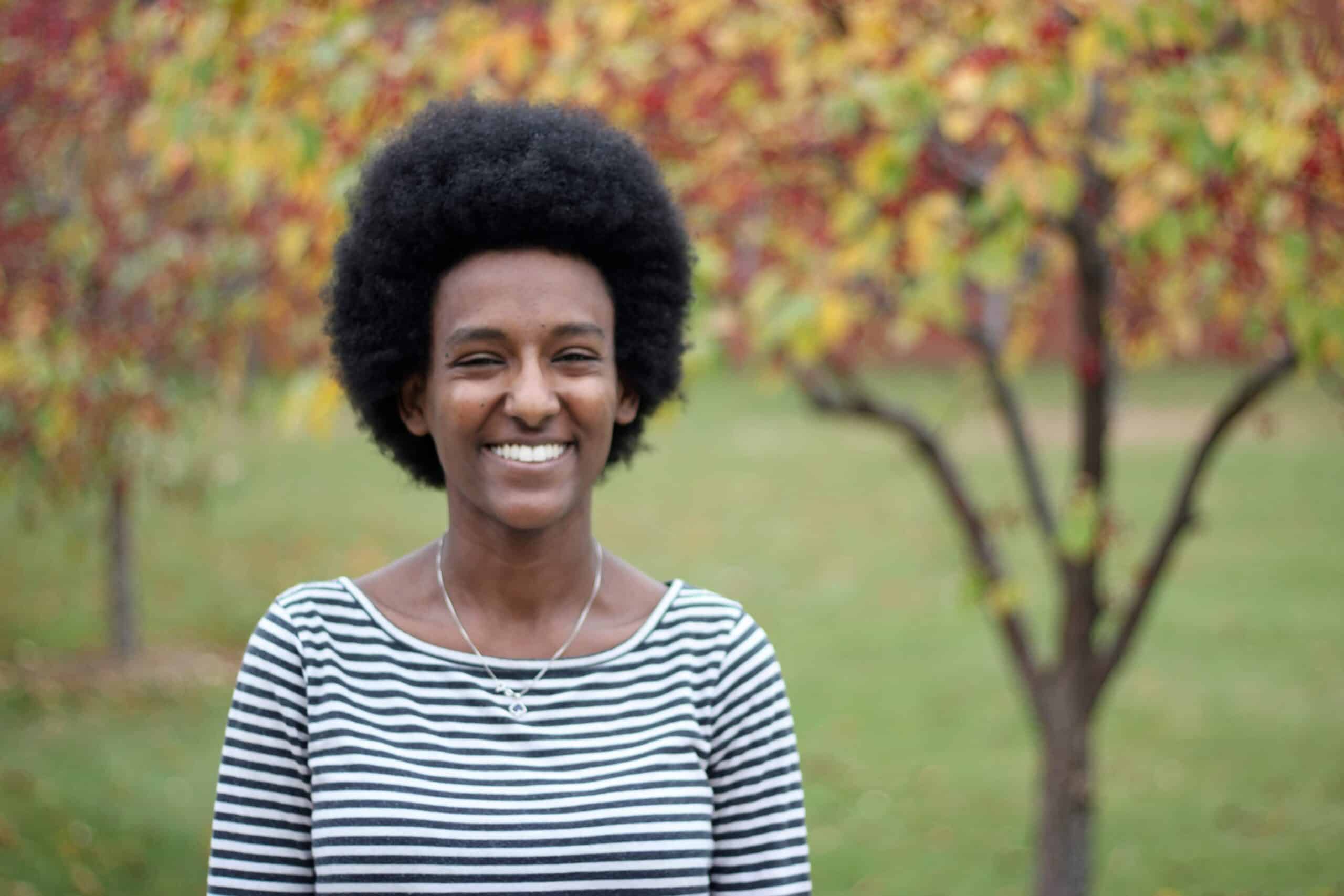 A portrait of a young woman with an afro smiling at the camera, fall foliage in the background.