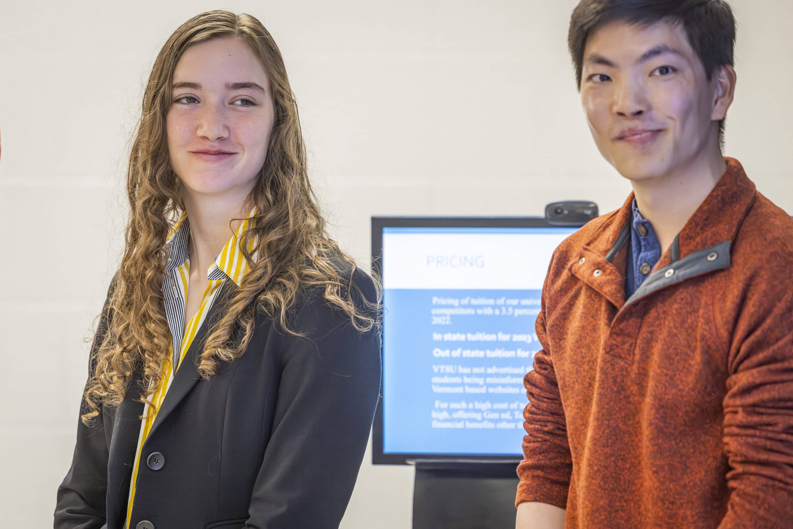 A woman in a suit and a man in a quarter-zip shirt stand at the front of a classroom giving a presentation