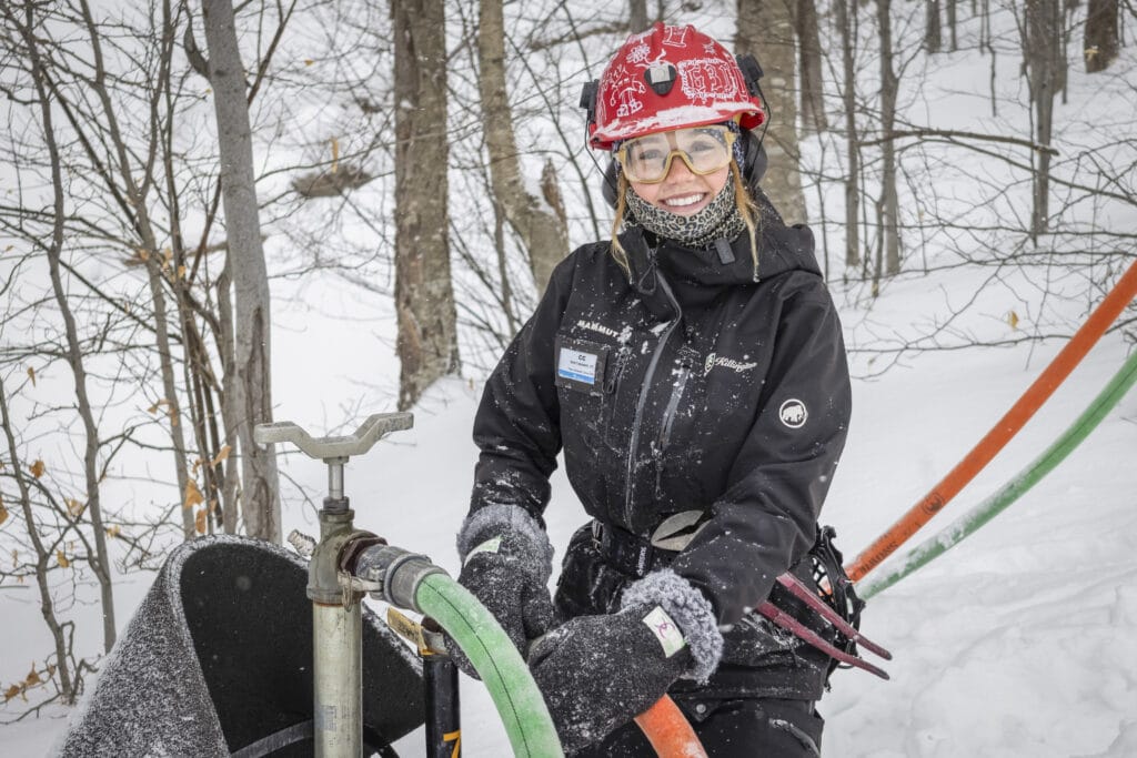 A student in a snow-jacket, snow-pants, and red hard hat is smiling at the camera while attending to a snowmaking machine.