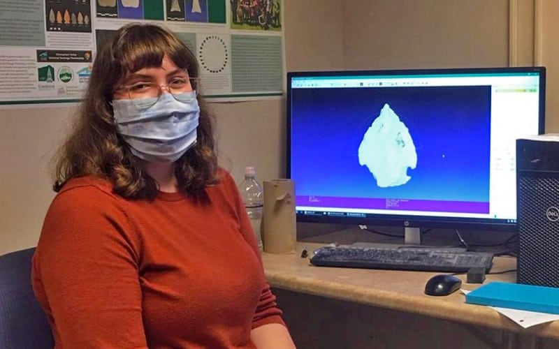 A woman with long brown hair sits at a desk looking at the camera wearing a hospital mask.